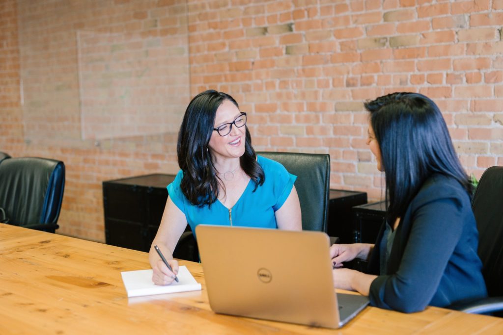 Two black haired business women talking at a wood conference table with a laptop while one smiles and writes on a notepad