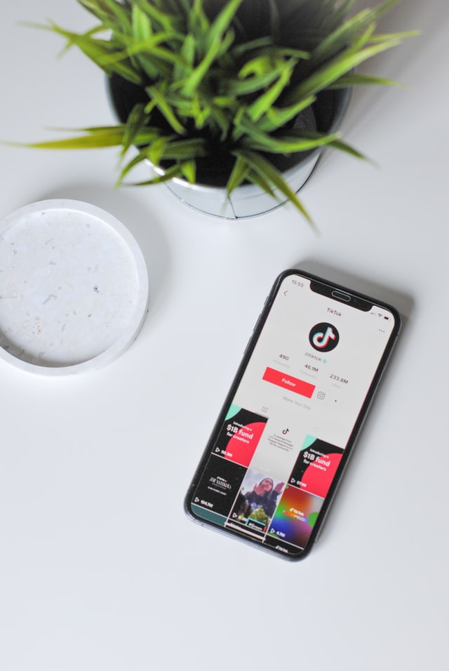 A smartphone showing the tik tok homepage on a white table with a plant on it