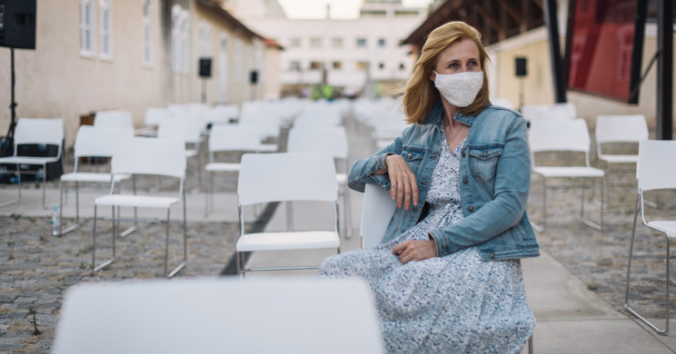 Blonde woman in a mask and a blue dress and denim jacked sitting in a chair with empty chairs around her outside