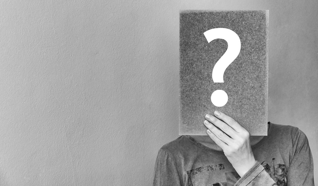 Black and white image of a person holding a paper in front of their face with a question mark on it