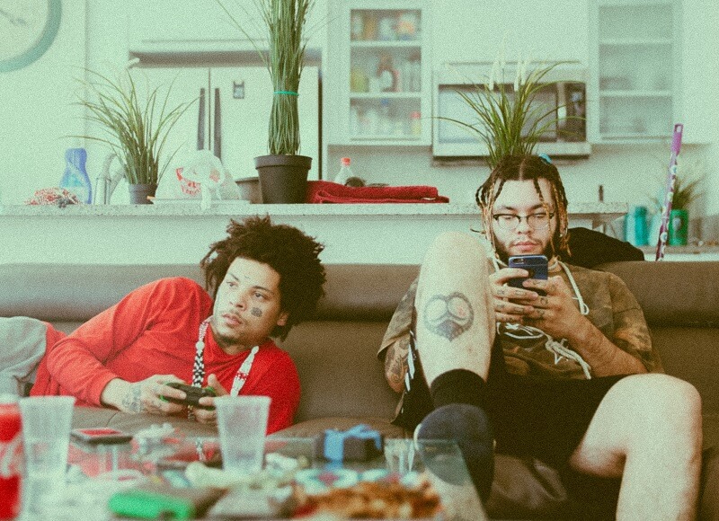 Two male friends relaxing on a couch one playing video games and the other on their smartphone
