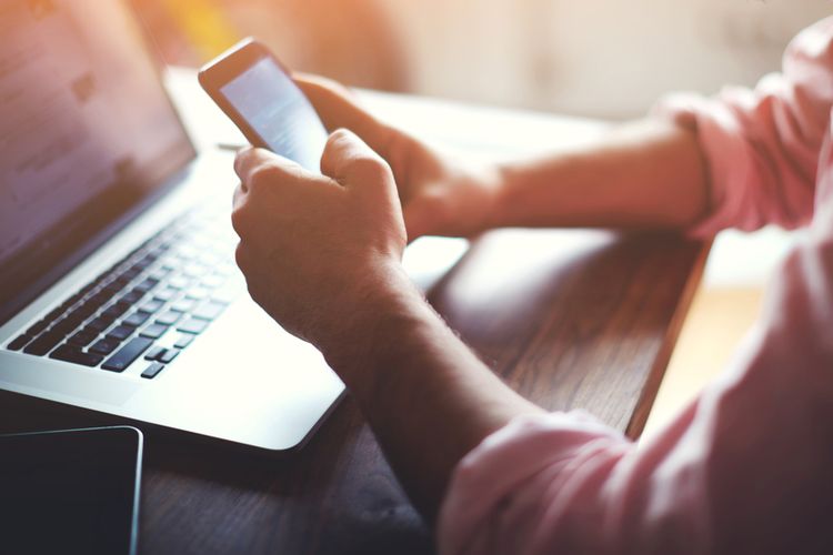 Man holding a smartphone in both hands above a laptop on a wooden table