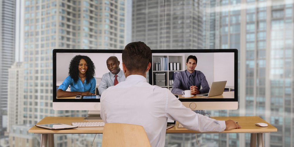 A man at a wood desk with two monitors that are displaying other coworkers in a video call