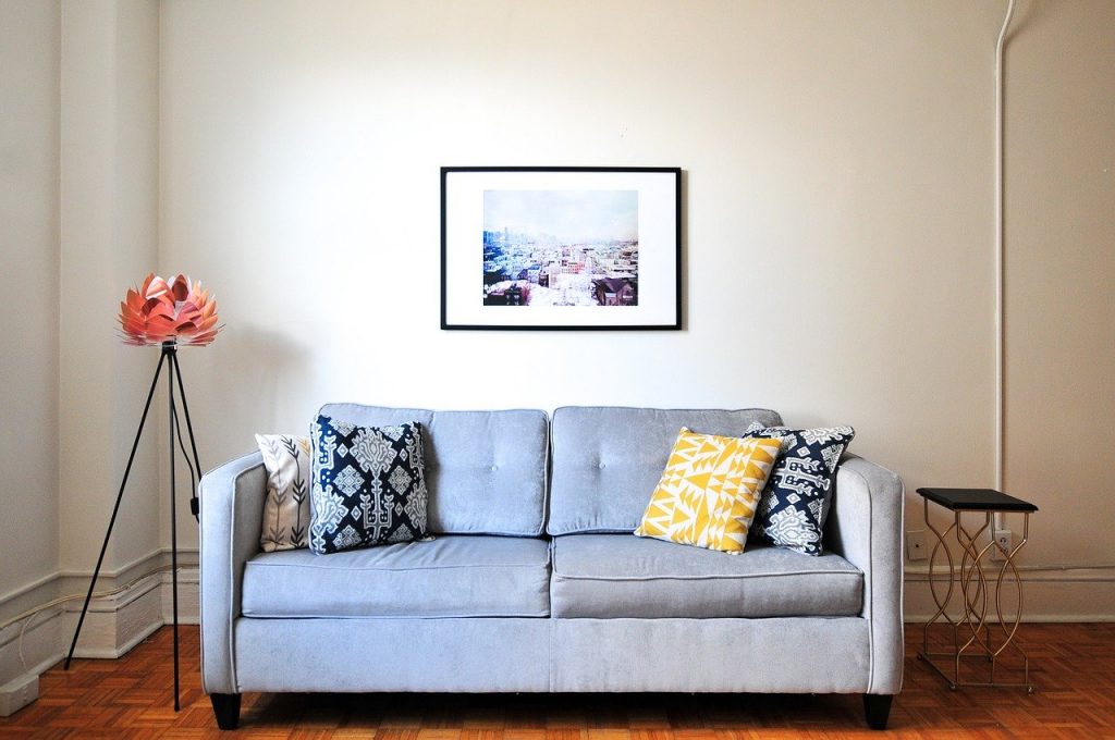 A gray couch in front of a white wall with a painting on a hardwood floor