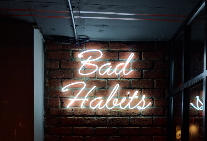 The text "Bad Habits" made by white neon lights illuminating a brick wall