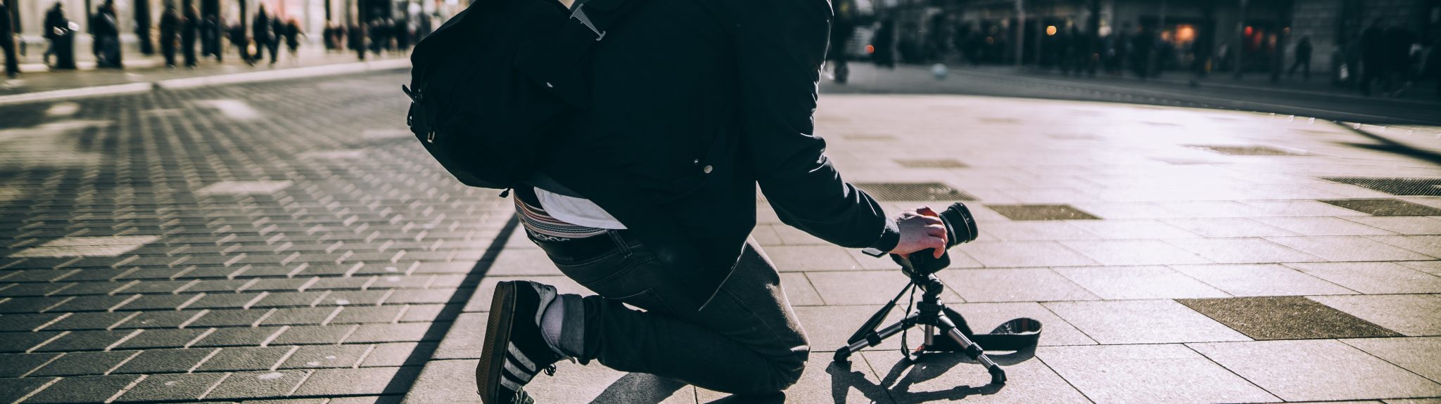 A person kneeling in a town square setting up a small camera on a tripod