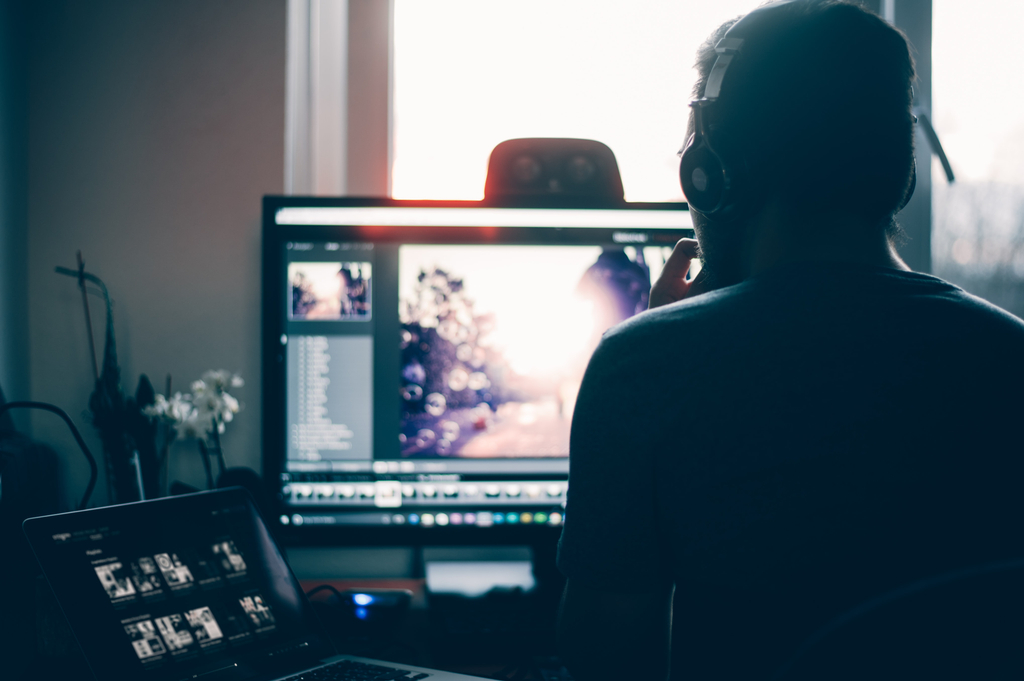 A man sitting at a desk with headphones with a laptop and monitor in front of him in a dark room with light coming through a window