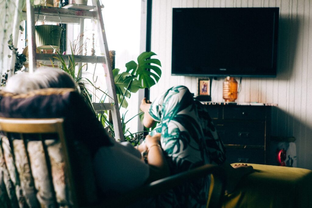 Woman lounging on a couch in an apartment room with a lot of plants looking at a wall mounted TV with nothing on it