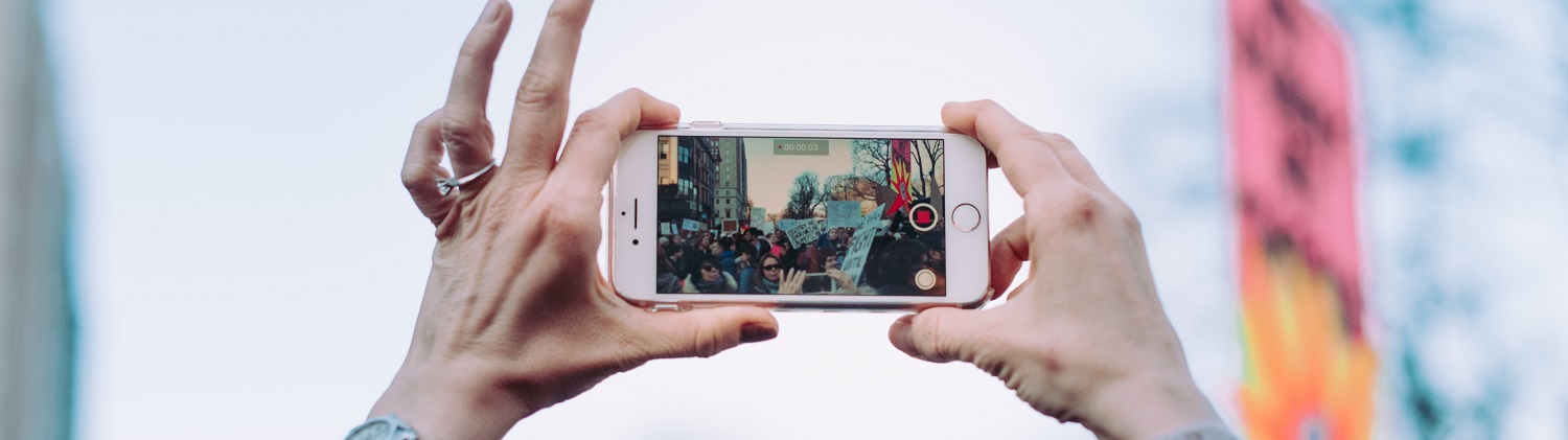 Close up of woman's hands holding an iPhone landscape recording a large group of people with signs