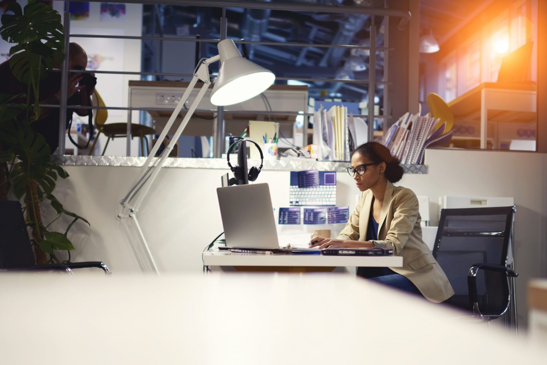 Sideview of a woman with black hair and glasses sitting at a cluttered desk working on a laptop in an office