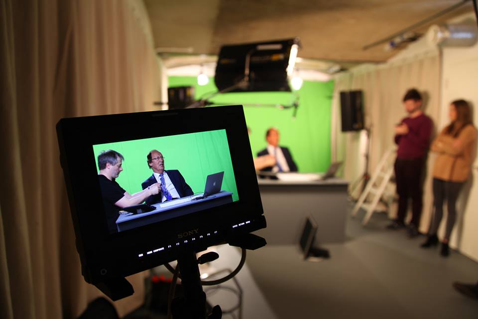 Close up of a small monitor of an actor being recorded at a desk with a green screen behind them as a technician works on a mic on the table