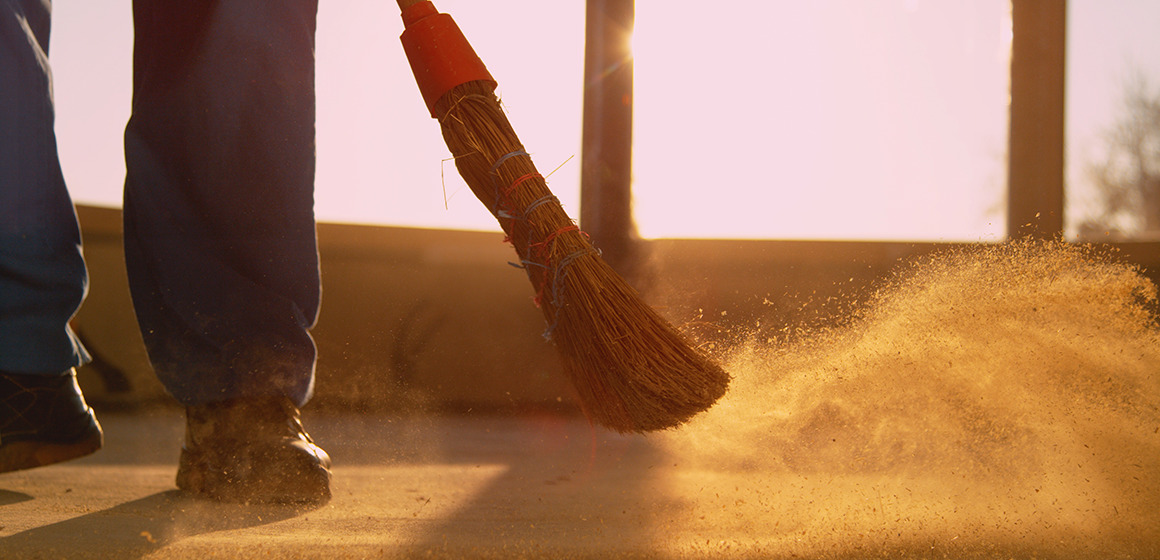 Image of a broom head and mans feet sweeping dirt up from a floor outside