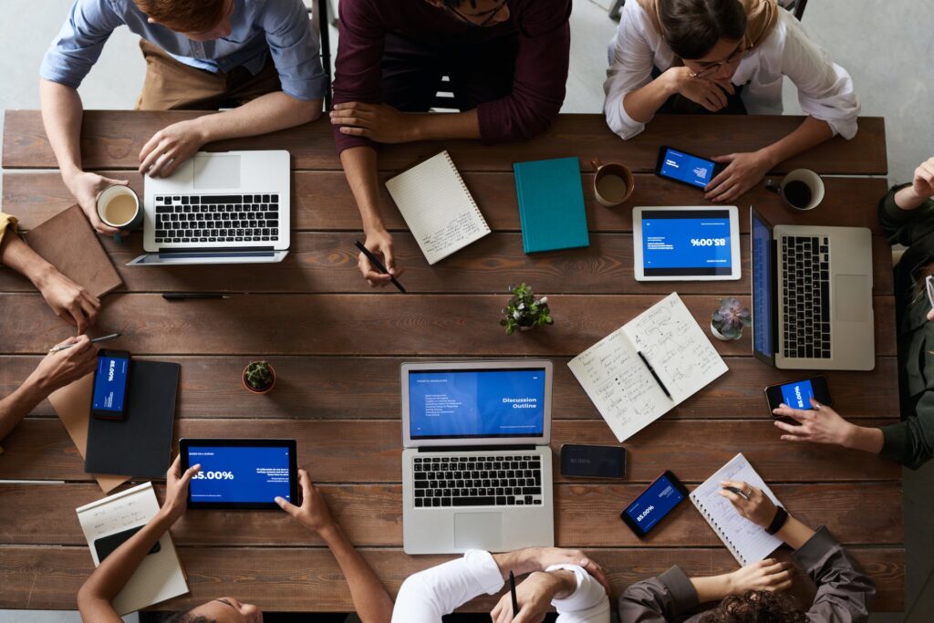 Aerial shot of a wooden picnic table with 8 individuals sitting around it with laptops, notebooks, smarthphones, tablets and coffees with two small plants in the middle