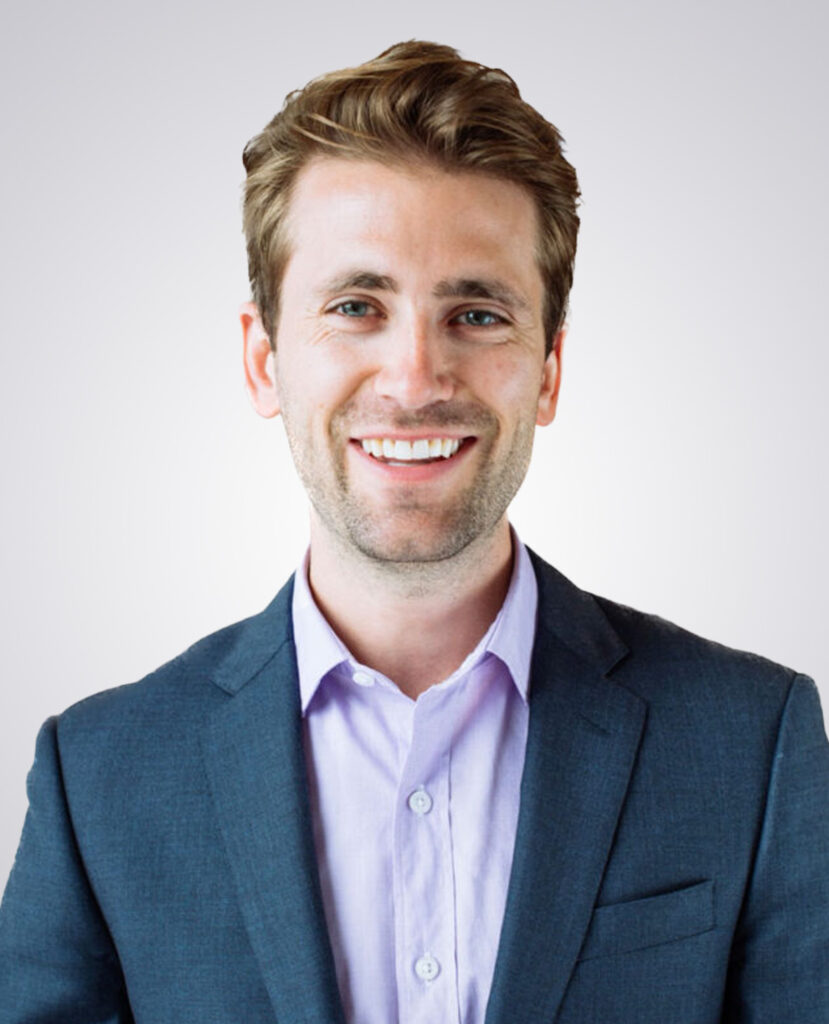 Professional headshot of man with blond hair smiling wearing a blue suit jacket and purple undershirt