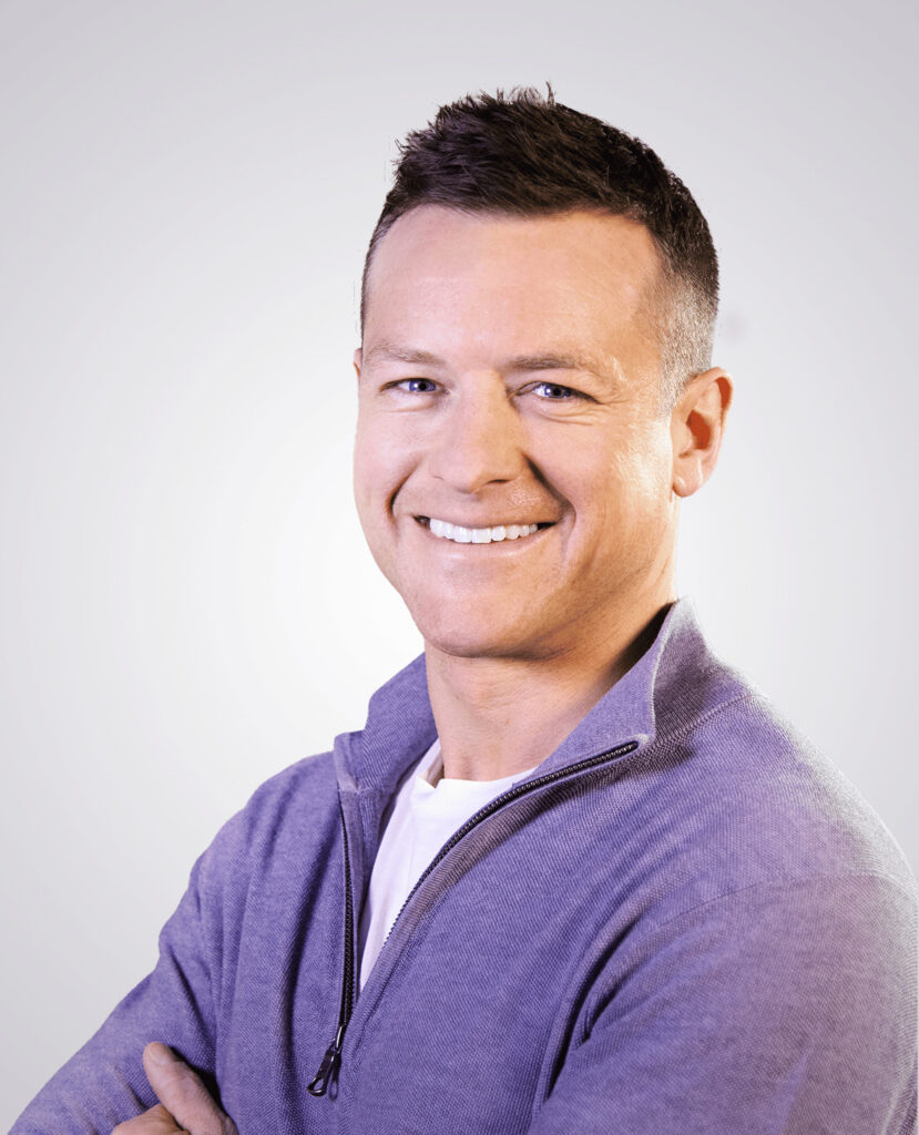 Professional headshot of man with short brown hair smiling and wearing a purple sweater