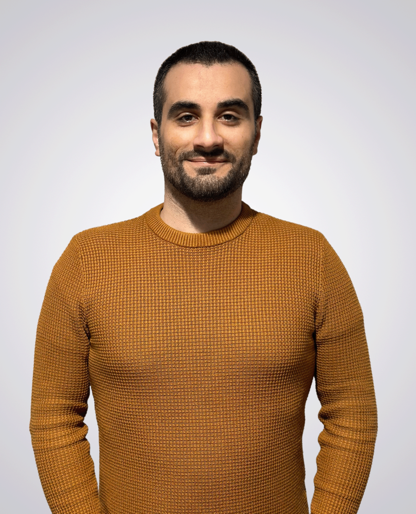 Professional headshot of a man with short black hair smiling wearing a orange sweater on a white background
