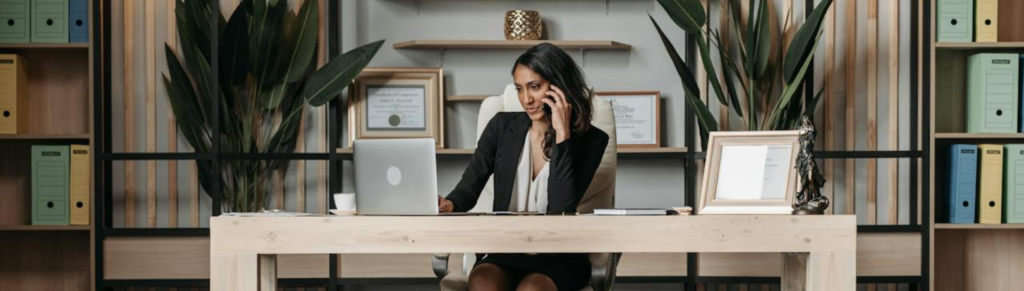 Image of women sitting at a light wooden desk on the phone while browsing a laptop with ferns and binders on the wall behind her