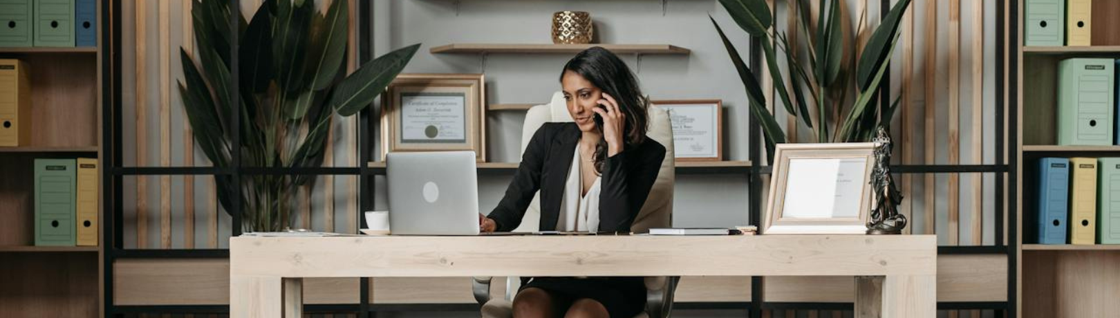Image of women sitting at a light wooden desk on the phone while browsing a laptop with ferns and binders on the wall behind her
