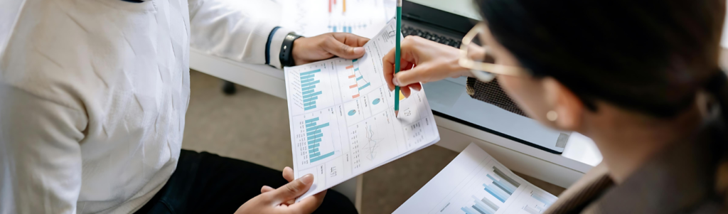 Woman pointing to a paper with a pencil that is held by another person that has graphic representations of statistics.